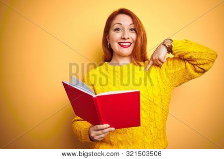 Young redhead teacher woman reading red book over yellow isolated background with surprise face pointing finger to himself
