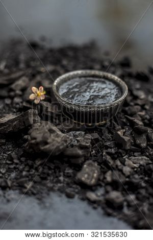 Close Up Of Activated Charcoal In A Glass Bowl On The Wooden Surface Along With Some Raw Powder Of C