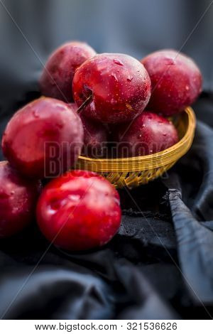 Raw Organic Red Ripe Plums In A Brown-colored Basket On Black Fabric On The Wooden Surface.