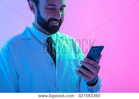Cropped portrait of a smiling young man doctor wearing uniform standing isolated over pink-blue haze backgroundm using mobile phone