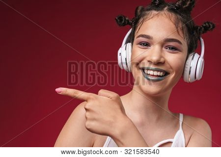 Image of optimistic punk girl with bizarre hairstyle pointing finger at copyspace while listening to music with headphones isolated over red background
