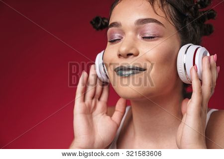 Image of optimistic punk girl with bizarre hairstyle and closed eyes smiling while listening to music with headphones isolated over red background