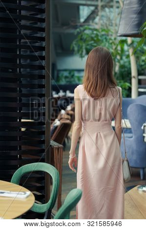 View From The Back Of A Slender Girl In A Long Pink Dress With Long Dark Hair In A Restaurant. Woman