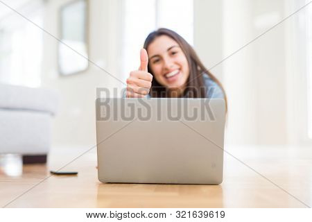 Beautiful young woman laying on the floor using laptop happy with big smile doing ok sign, thumb up with fingers, excellent sign