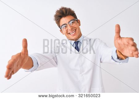 Young handsome sciencist man wearing glasses and coat over isolated white background looking at the camera smiling with open arms for hug. Cheerful expression embracing happiness.