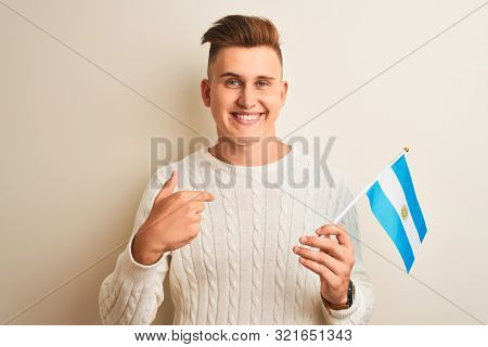 Young handsome man holding Argentinian Argentina flag over isolated white background with surprise face pointing finger to himself