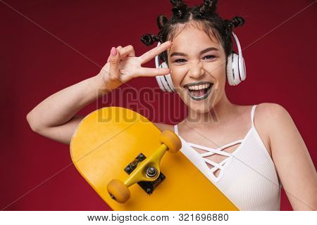 Image of optimistic punk girl with bizarre hairstyle and dark lipstick holding skateboard while listening to music with headphones isolated over red background