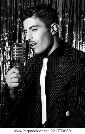 Attractive Latino Man Singing In Front Of Vintage Microphone In Nightclub With Silver Curtain In Bac
