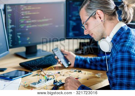 Developer Is Connecting Breadboard To Microcontroller. Man Is Holding Smartphone With Program Code S