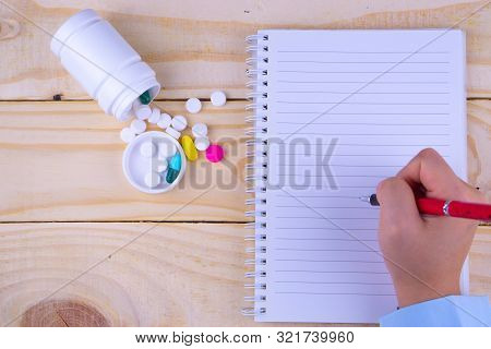 Top View Of Woman Hands Holding Pen To Writing A Book With White Bottle And Heap Of Pill On Wooden T