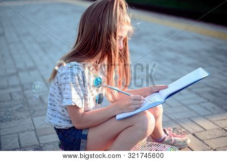 Beautiful Little Girl Sitting On A Skateboard And Writing In A Tetrad. Outdoor Lifestyle