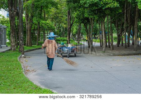 An Asian Woman Is Sweeping Dry Leaves By The Road In The Outdoor Garden. Cleaning Concept.