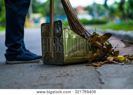 An Asian Woman Is Sweeping Dry Leaves By The Road In The Outdoor Garden. Cleaning Concept.