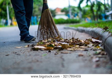 An Asian Woman Is Sweeping Dry Leaves By The Road In The Outdoor Garden. Cleaning Concept.