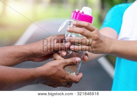 Hands Of Senior Man Holding Bottle Of Fresh Water For His Wife While Workout In The Park. Healthy Li