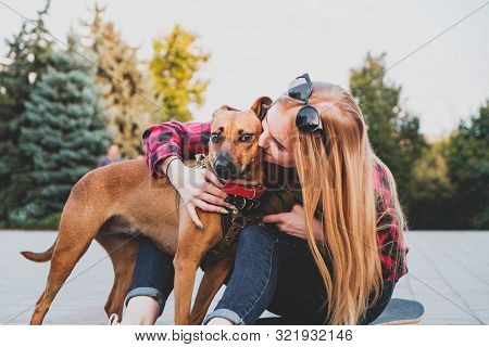 Teenage Skater Girl Kissing Her Dog In Urban Environment. Mixed Breed Dog Stands Next To Young Woman