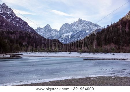 Beautiful View At Sunset Of The Peaceful Lake Fucine Tarvisio, Italy With Green Forest And Snowy Mou