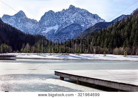 Beautiful View At Sunset Of The Peaceful Lake Fucine Tarvisio, Italy With Green Forest And Snowy Mou