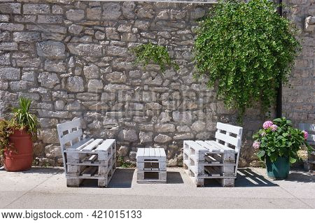 Benches Are Situated Beneath Greenery In Front Of An Historical Stone Wall Between Two Flower Pots