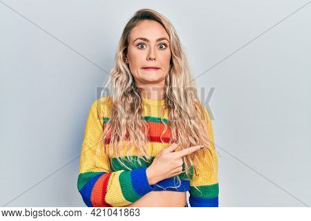 Beautiful young blonde woman wearing colored sweater pointing aside worried and nervous with forefinger, concerned and surprised expression 