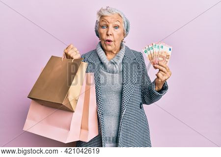 Senior grey-haired woman holding shopping bags and euros banknotes in shock face, looking skeptical and sarcastic, surprised with open mouth 