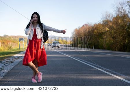 A Young Beautiful Brunette In A Red Dress Hitchhiking And Calling On The Phone. In The Background, T