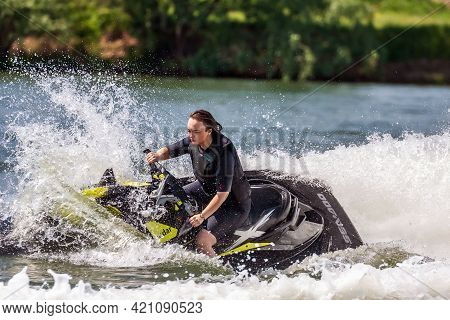 Krasnodar, Russia - July 25, 2020: Young Caucaisian Woman Drives Jet Ski Splashing At Sunset By Rive