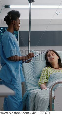 Afro American Asisstant Checking Sick Woman While Sitting In Bed In Hospital Ward. Medical Nurse Wri