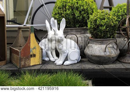 Two Ceramic Hares Among Boxwood Bushes And Garden Grass. Trolley On Wheels And Miscellaneous Gardeni