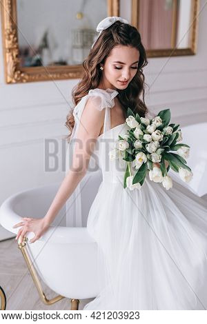 Beautiful Bride With A Wedding Bouquet Sitting On Sofa Indoors In White Studio Interior Like At Home