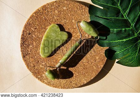 Jade Gua Sha Scraper And Face Roller Massager On A Cork Round Stand With A Monstera Leaf. Hard Light