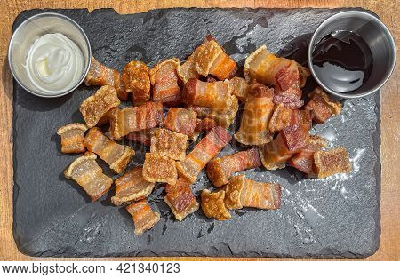 Plate Of Fried Torreznos Or Fried Rashers Seen From Above On A Black Slate Plate And Two Jars With M