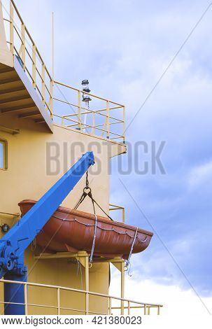 Low Angle View Of Orange Lifeboat And Crane Boom On Deck Of Cargo Ship Against Rain Clouds In Vertic