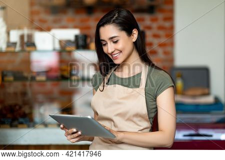 Young Female Owner Of Modern Cafe In Apron Holding Digital Tablet And Standing With Shopwindow On Ba