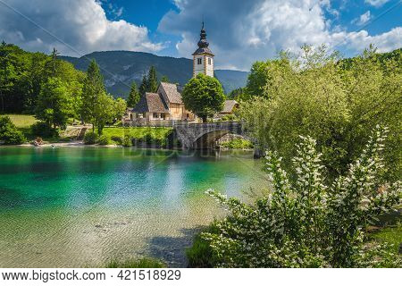 Flowery Lake Shore And Old Stone Bridge Over The Lake Bohinj. Traditional Old Church On The Shore Of