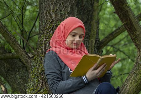 Cute Muslim Student Reading A Book While Sitting On The Tree Branch In The Park. Modern And Fashiona