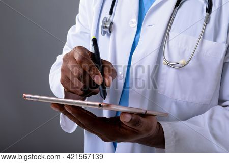 Mid section of african american male doctor using graphic tablet against grey background. healthcare and medical professionalism concept