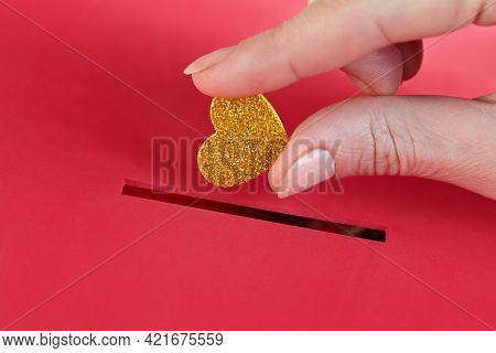 Female Hand Puts Gold Heart Into Slot Of Red Donation Box. Concept Of Sincere Devotion To Faith. Con