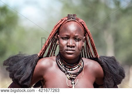 OPUWO, OTJIWARONGO REGION NAMIBIA - MAY 4, 2021: Young unidentified Himba woman with the typical necklace and hairstyle in Himba living museum during a appearance in front of tourists