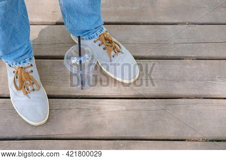 Legs Fragment Standing On A Wooden Planks Surface In Blue Jeans With Empty Tall Plastic Glass Betwee