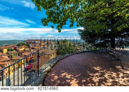 Bench under trees on a viewpoint overlooking small town of Diano d'Alba under beautiful sky in Piedmont, Northern Italy.