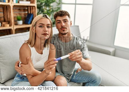 Young caucasian couple with serious expression holding pregnacy test at home.