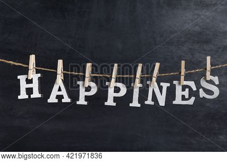 Word Happines Held On A Clothespin On A Rope Against Black Background. Inscription From White Letter