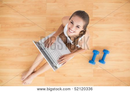 Smiling sporty brunette using a laptop and sitting next to dumbbells on the floor
