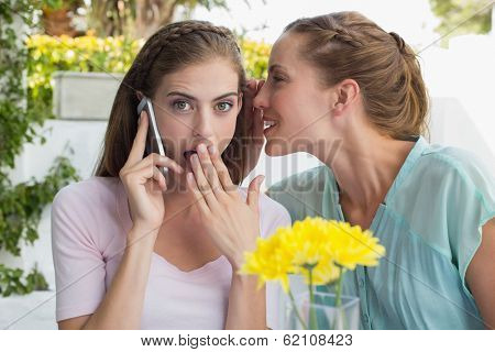 Young woman whispering secret into a friends ear while shes on call at cafe