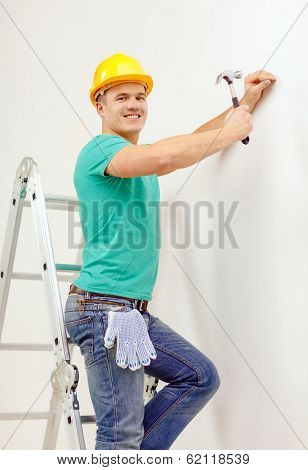 reapir, building and home renovation concept - smiling man in yellow protective helmet hammering nail in wall