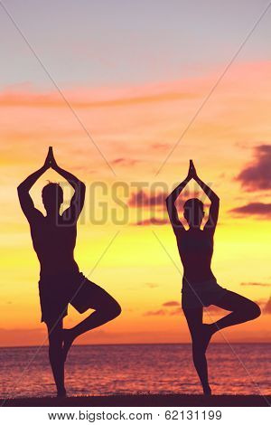 Yoga couple training in sunset in tree pose meditating outdoors by beach ocean sea. Man and woman working out training in serene ocean landscape. Silhouette against sunset