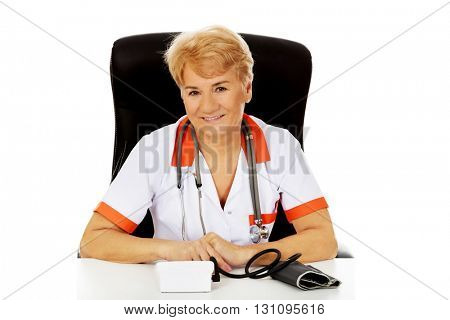 Smile elderly female doctor or nurse sitting behind the desk with bloog preasure gauge