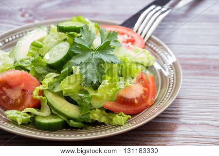 Diadonally a salad of lettuce, tomato, cucumber, avocado and cutlery on wood background. Healthy fresh salad for lunch. Horizontal. Daylight. Close.