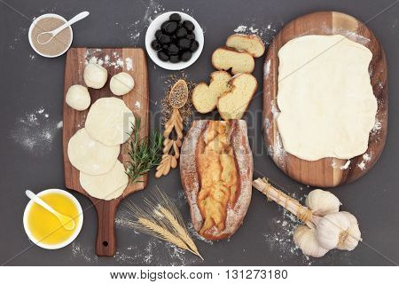 Baking ingredients with rustic bread loaf, dough, yeast, black olives, olive oil, garlic, herb and rye grain in a love spoon on maple wood over grey.